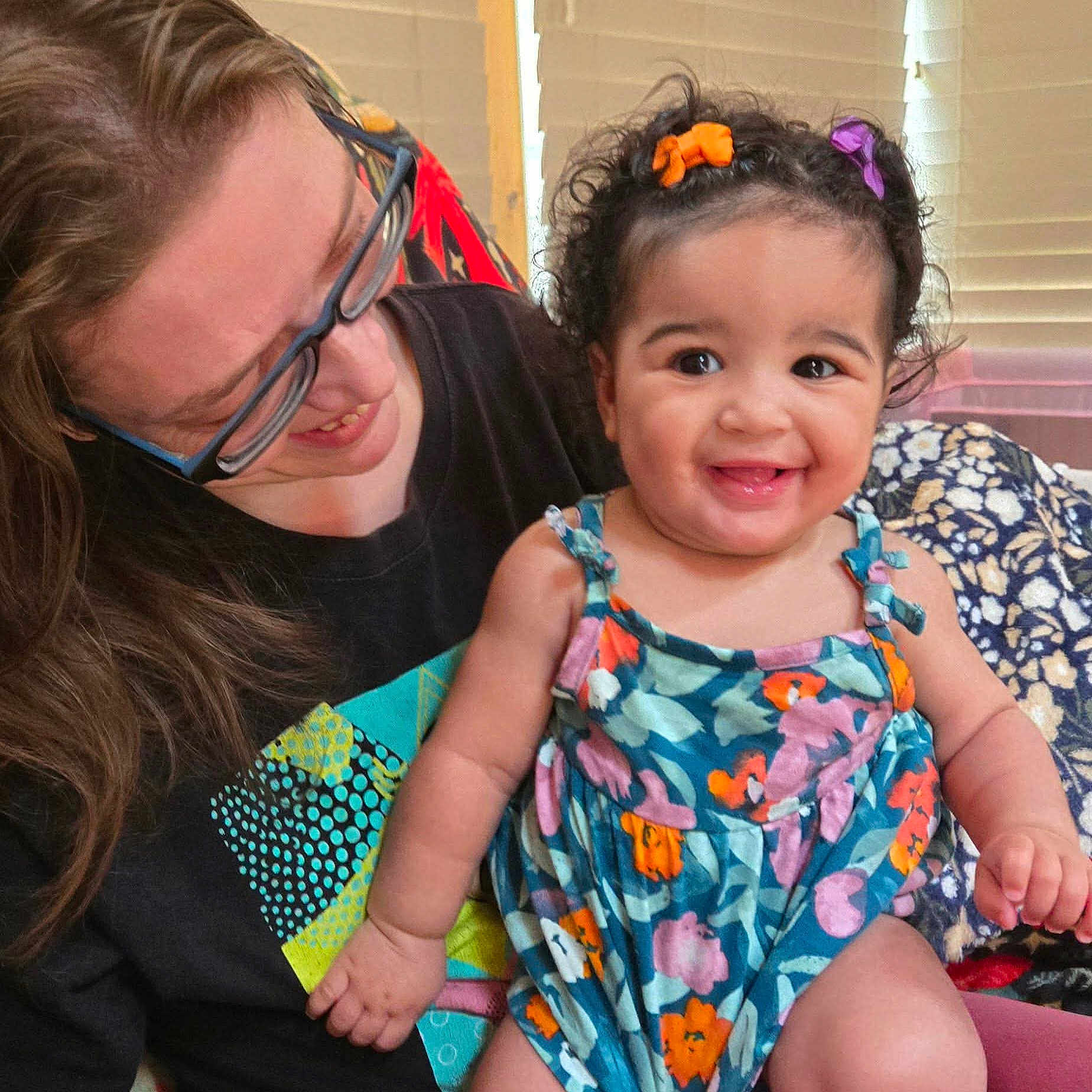 Nala Ann is registered to the contest to win money with this photo: baby, child, adult, glasses, hair_clip, floral_dress, smile, curly_hair, indoor, blanket, patterned_clothing, person, happy, holding, seated, window_blinds, family, cozy, portrait, candid