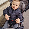 baby_stroller, baby_toy, casual_clothing, child, cute, daylight, happy, jacket, outdoor, person, portrait, seatbelt, sidewalk, smiling, stroller, toddler, transportation, urban, walking_path, young_child