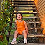 Dakohta is registered to the contest to win money with this photo: child, toddler, orange_outfit, sneakers, stairs, metal_stairs, outdoor, greenery, vines, smiling, happy, sunlight, rustic, brick_wall, grass, casual, sitting, nature, daylight, portrait