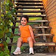 Dakohta is registered to the contest to win money with this photo: child, toddler, orange_outfit, sneakers, stairs, metal_stairs, brick_wall, green_leaves, outdoor, smiling, happy, sitting, sunlight, nature, plant, grass, portrait, daylight, person, casual