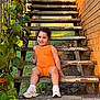 child, toddler, orange_outfit, sneakers, stairs, metal_stairs, brick_wall, green_leaves, outdoor, smiling, happy, sitting, sunlight, nature, plant, grass, portrait, daylight, person, casual