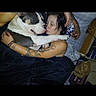 dog, person, tattoo, bed, blanket, cuddle, indoor, affection, black_hair, casual, resting, snuggle, comfort, pet, cozy, relaxation, human, animal, love, home