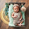 baby, sleeping, infant, wooden_bed, knitwear, pom_pom_hat, green_padding, woven_mat, wooden_floor, plant_leaves, peaceful, indoor, portrait, cozy, resting, newborn, soft_texture, cute, child, hat