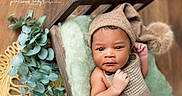 Moneii James is registered to the contest to win money with this photo: baby, infant, hat, pom_pom, wooden_bed, blanket, greenery, plant, fuzzy_texture, cozy, portrait, cute, newborn, child, indoors, soft_lighting, brown, beige, serious_expression, photography
