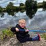 Jack-junior is registered to the contest to win money with this photo: child, boy, pond, water, fishing_rod, rock, grass, cloudy_sky, reflection, black_vest, rain_boots, outdoor, nature, smiling, sitting, playful, greenery, casual_clothing, daytime, recreation