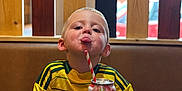 Jack-junior is registered to the contest to win money with this photo: child, boy, tongue_out, yellow_jersey, sitting, table, can, strawberry_soda, straw, wooden_slats, background, indoor, playful, casual, drink, short_hair, hands_on_table, smiling, face, portrait