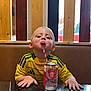 child, boy, tongue_out, yellow_jersey, sitting, table, can, strawberry_soda, straw, wooden_slats, background, indoor, playful, casual, drink, short_hair, hands_on_table, smiling, face, portrait