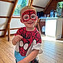 child, boy, face_paint, spiderman, costume, smiling, indoors, wooden_floor, window, natural_light, shirt, shorts, happy, person, playful, home, wooden_wall, casual_clothing, portrait, standing