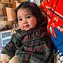 baby, child, cute, plaid_dress, bow, black_hair, orange_sweater, cartoon_character, kitchen, indoor, person_holding, warm_lighting, smiling, chubby_cheeks, long_sleeves, cozy, portrait, happy, infant, close_up