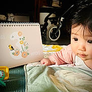 Himéa participe au concours pour gagner de l'argent avec cette photo : baby, infant, child, face, eyes, toy, quilt, blanket, calendar, age_marker, pink_clothing, floor, stroller, wheel, flower_illustration, bird_illustration, hand, curious, indoor, closeup