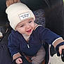 baby, child, smiling, beanie, hat, pompon, jacket, stroller, outdoor, happy, cute, infant, person, clothing, cold_weather, sitting, playful, young, expression, portrait