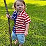 Jackson is registered to the contest to win money with this photo: toddler, child, barefoot, grass, stick, outdoor, summer, greenery, shirt, shorts, flag_pattern, blue_eyes, curly_hair, nature, park, sunlight, casual_clothing, standing, young_child, daytime