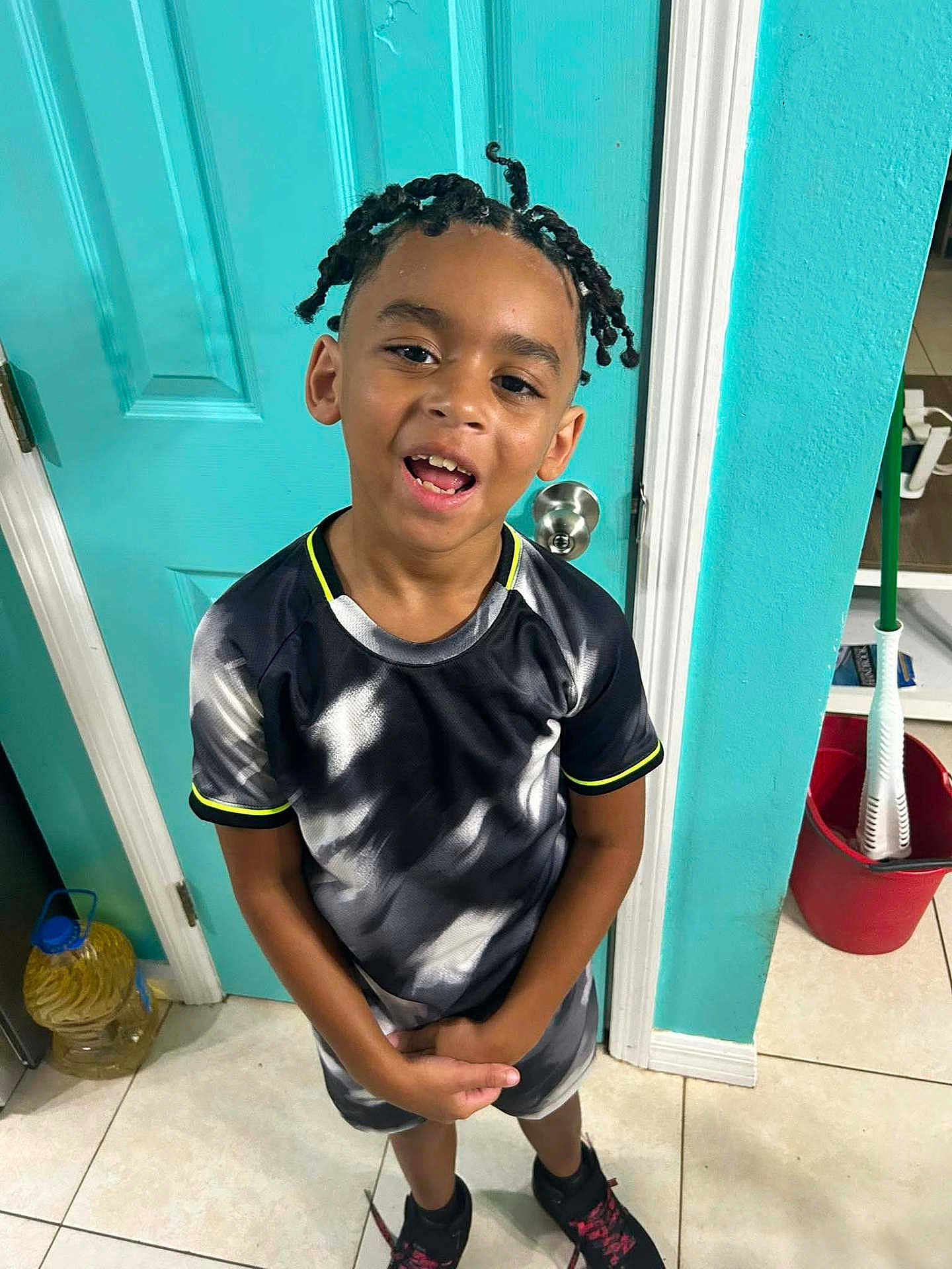Nicholas is registered to the contest to win money with this photo: child, boy, smiling, braided_hair, tshirt, shorts, sneakers, indoors, door, tile_floor, bucket, mop, cleaning_supplies, playful, casual, portrait, standing, happy, young, home