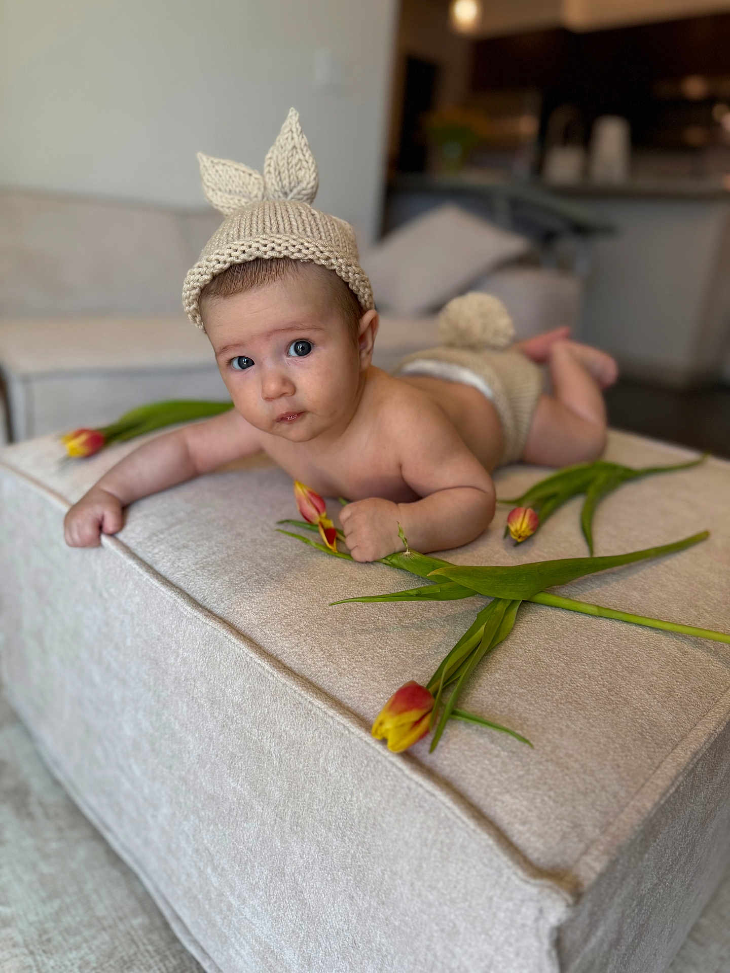 Arthur joined the competition — help win amazing prizes! baby, infant, knitted_hat, bunny_ears, tulips, flower, ottoman, beige, indoor, living_room, cute, child, portrait, soft_lighting, couch, home_decor, laying_down, curious, expression, hand