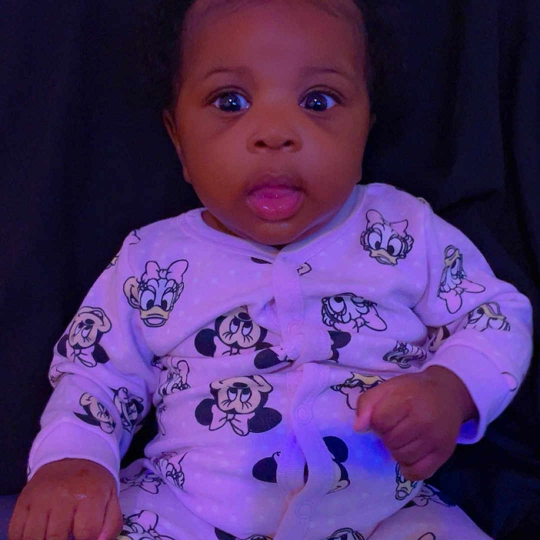 Sariyah is registered to the contest to win money with this photo: baby, child, onesie, mickey_mouse, minnie_mouse, infant, cute, tongue_out, sitting, purple_light, indoor, portrait, person, clothing, face, young_child, expression, dark_background, soft_lighting, adorable