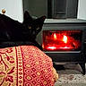 black_cat, cat, feline, pet, animal, indoors, living_room, wood_stove, fireplace, fire, flames, cozy, warmth, blanket, patterned_blanket, red, gold, furniture, floor, resting