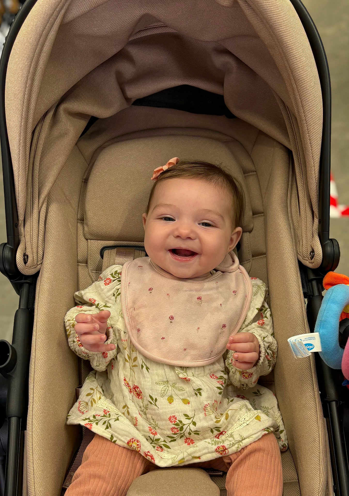 Emila joined the competition — help win amazing prizes! baby, smiling, stroller, pink_bib, floral_dress, infant, child, toy, happy, seat, cute, clothing, headwear, indoors, person, legs, fabric, comfort, plaything, portrait
