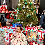 Amelia joined the competition — help win amazing prizes! baby, child, christmas_tree, christmas_lights, christmas_gifts, gift_bags, pajamas, pink_clothing, holiday_decor, ornaments, green_ornaments, silver_ornaments, smiling, sitting, floor, wooden_floor, indoor, festive, holiday, happy