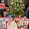 baby, child, christmas_tree, christmas_lights, christmas_gifts, gift_bags, pajamas, pink_clothing, holiday_decor, ornaments, green_ornaments, silver_ornaments, smiling, sitting, floor, wooden_floor, indoor, festive, holiday, happy