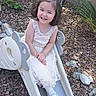 Hana joined the competition — help win amazing prizes! child, girl, toddler, smiling, happy, portrait, outdoor, playground, slide, dress, white_dress, hair_bow, long_hair, sitting, garden, rocks, plants, backyard, sunlight, playtime