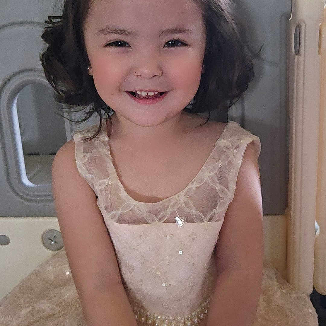 Hana joined the competition — help win amazing prizes! child, girl, smiling, portrait, indoors, dress, princess_dress, bow, hair, brown_hair, seated, hospital_bed, medical_room, sparkly, formal_wear, happy, cute, face, person, photography