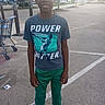 baseball_graphic, basketball_graphic, boy, casual_clothing, child, daylight, green_pants, outdoor, parking_lot, parking_spaces, pavement, portrait, shopping_cart, smiling, sneakers, sportswear, standing, store, t_shirt, trees