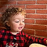 child, toddler, curly_hair, red_onesie, checkered_pattern, reindeer_design, brick_wall, indoor, hand, cute, portrait, smiling, baby, face, person, young_child, warm_lighting, cozy, clothing, curious