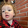 child, toddler, curly_hair, red_clothing, checkered_pattern, face, expression, indoor, brick_wall, background, clothing, person, portrait, close_up, young_child, casual, curious, head_and_shoulders, indoor_lighting