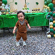Logan is registered to the contest to win money with this photo: child, toddler, birthday, party, cake, cupcakes, balloons, dessert_table, outdoor, smiling, standing, brown_outfit, scarf, sneakers, green_decorations, jungle_theme, birthday_cake, celebration, gravel, portrait