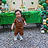 child, toddler, birthday, party, cake, cupcakes, balloons, dessert_table, outdoor, smiling, standing, brown_outfit, scarf, sneakers, green_decorations, jungle_theme, birthday_cake, celebration, gravel, portrait