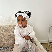 Lilena a rejoint le concours — aidez-le/la à gagner de superbes lots ! toddler, child, pacifier, headband, white_bow, curly_hair, white_blouse, beige_pants, sitting, couch, fluffy_pillow, plastic_bag, indoor, cozy, looking_away, curly_hair_buns, floor, side_table, black_and_white_shoes, casual_clothing