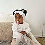 toddler, child, pacifier, headband, white_bow, curly_hair, white_blouse, beige_pants, sitting, couch, fluffy_pillow, plastic_bag, indoor, cozy, looking_away, curly_hair_buns, floor, side_table, black_and_white_shoes, casual_clothing