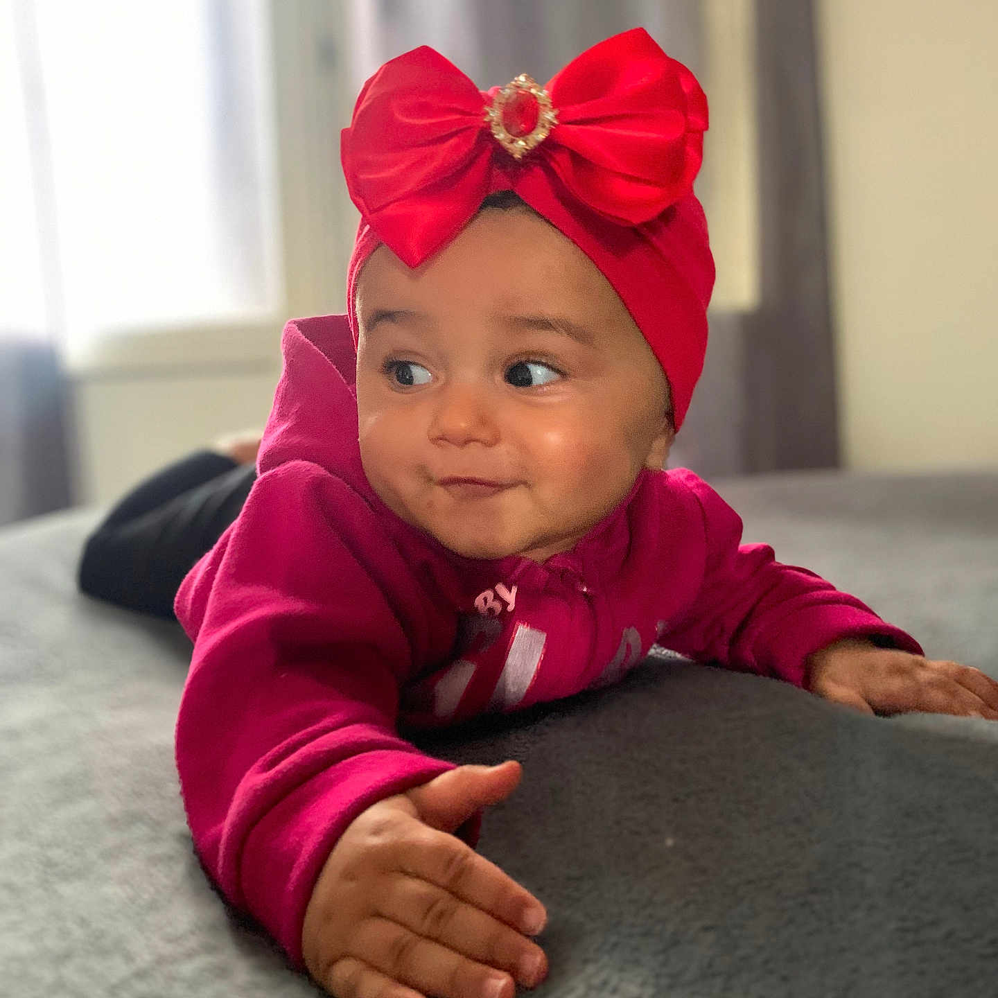 Lilena participe au concours pour gagner de l'argent avec cette photo : baby, bed, child, clothing, cozy, cute, expression, face, gray_bedspread, hand, headband, indoor, infant, magenta_hoodie, natural_light, person, portrait, red_bow, smile, window