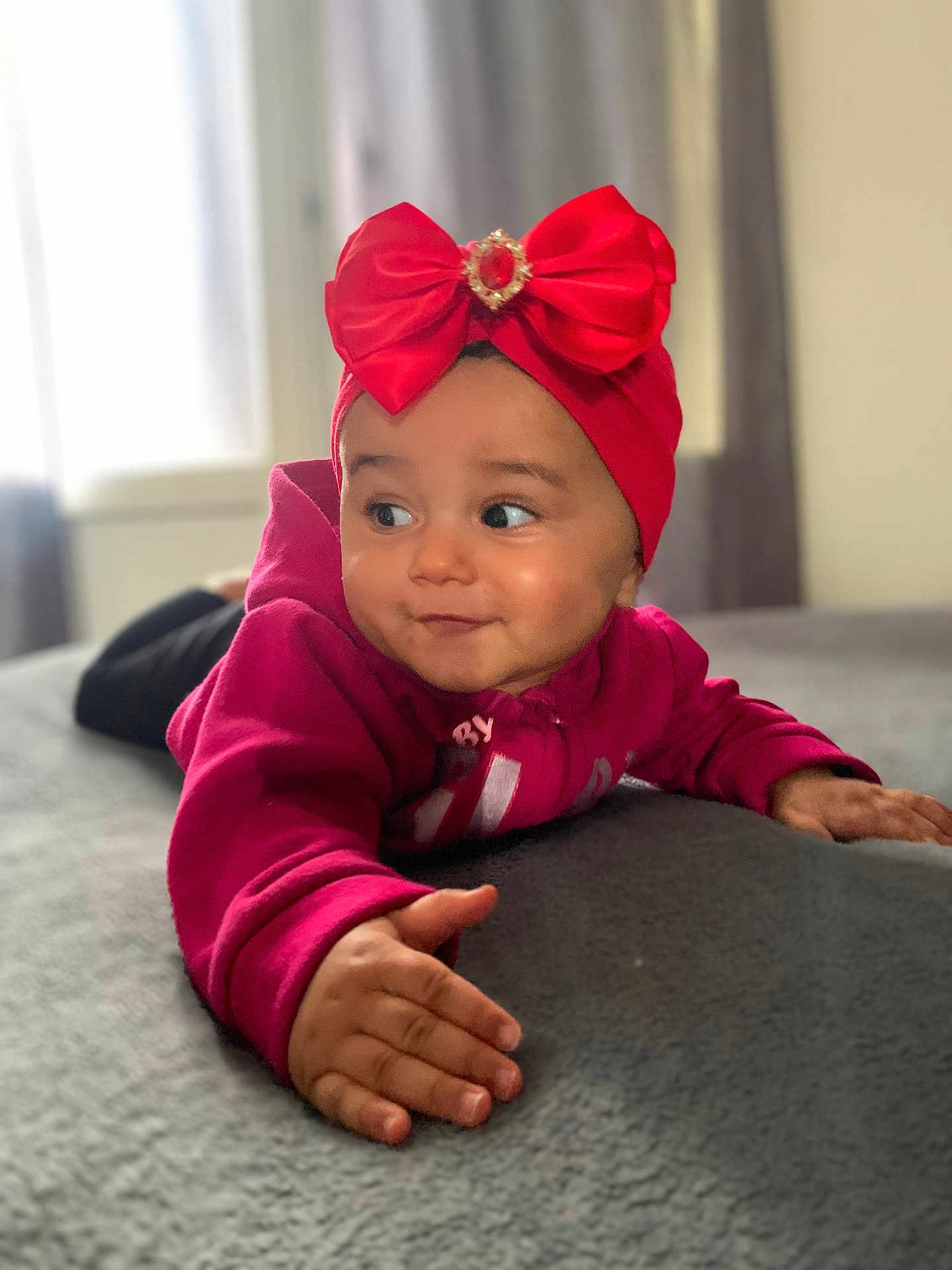 Lilena participe au concours pour gagner de l'argent avec cette photo : baby, child, red_bow, headband, magenta_hoodie, bed, gray_bedspread, indoor, smile, face, hand, cute, portrait, person, infant, expression, natural_light, window, cozy, clothing