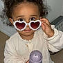 Lilena participe au concours pour gagner de l'argent avec cette photo : child, curly_hair, sunglasses, heart_shaped, flower_decor, holding, sippy_cup, indoor, curious_look, cute, white_shirt, long_sleeves, brown_skin, big_eyes, standing, floor, radiator, casual_clothing, portrait, close_up