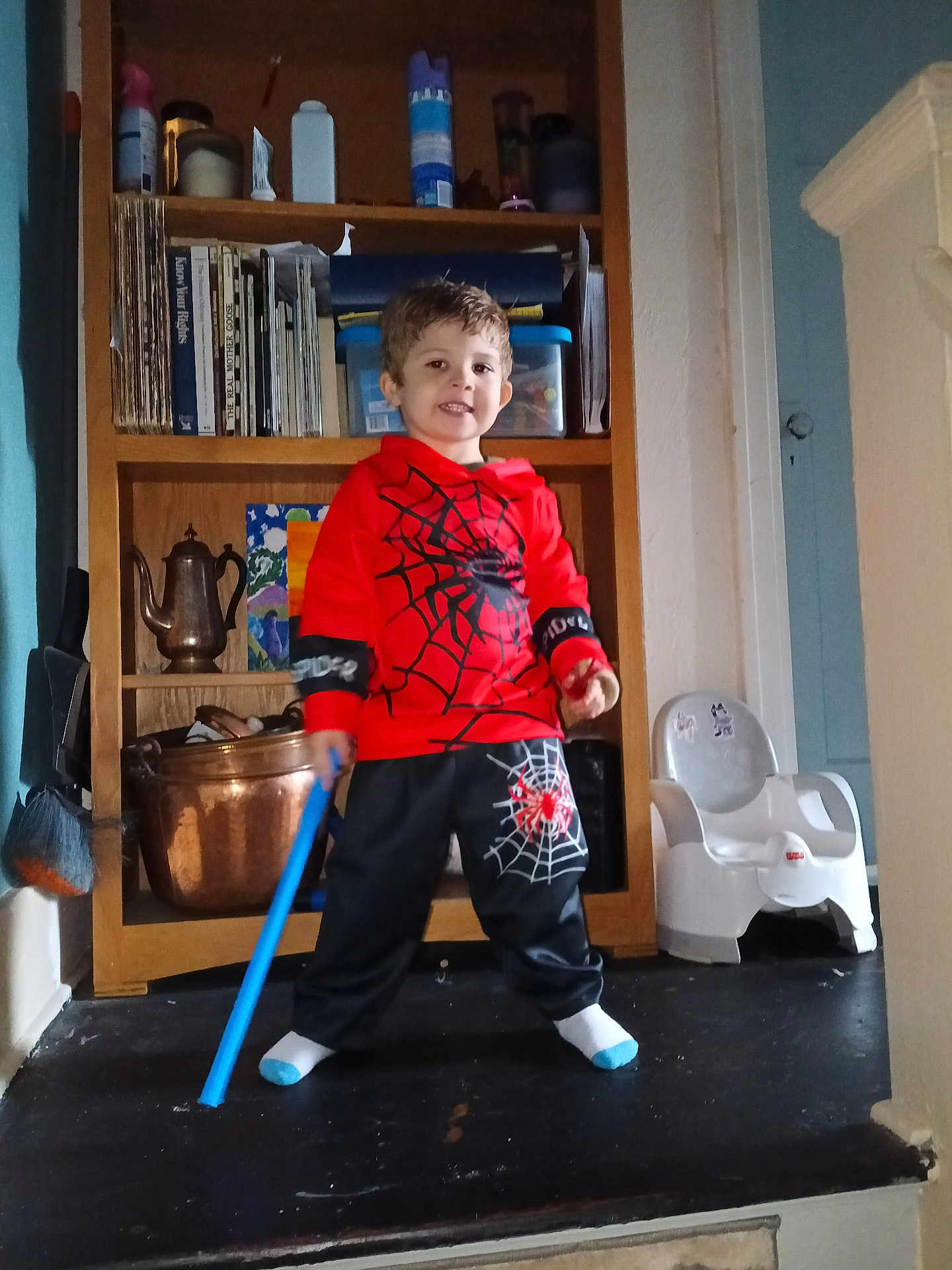 Bentley joined the competition — help win amazing prizes! child, toddler, spiderman_outfit, red_clothing, black_pants, blue_toy, toy_stick, smiling, indoor, wooden_shelf, books, potty_chair, floor, household_items, copper_pot, face, person, white_socks, short_hair, playful