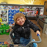 Bentley is registered to the contest to win money with this photo: aisle, blonde_hair, blue_shoes, canned_food, casual, child, clothing, grocery_store, happy, holding, indoor, jacket, person, plastic_bottles, produce, shelves, shopping_cart, smiling, store_interior, supermarket