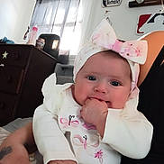 Summer is registered to the contest to win money with this photo: baby, infant, child, headband, bow, hand, face, eyes, person, sleeve, clothing, furniture, curtain, window, dresser, baseball, decor, indoor, holding, adult