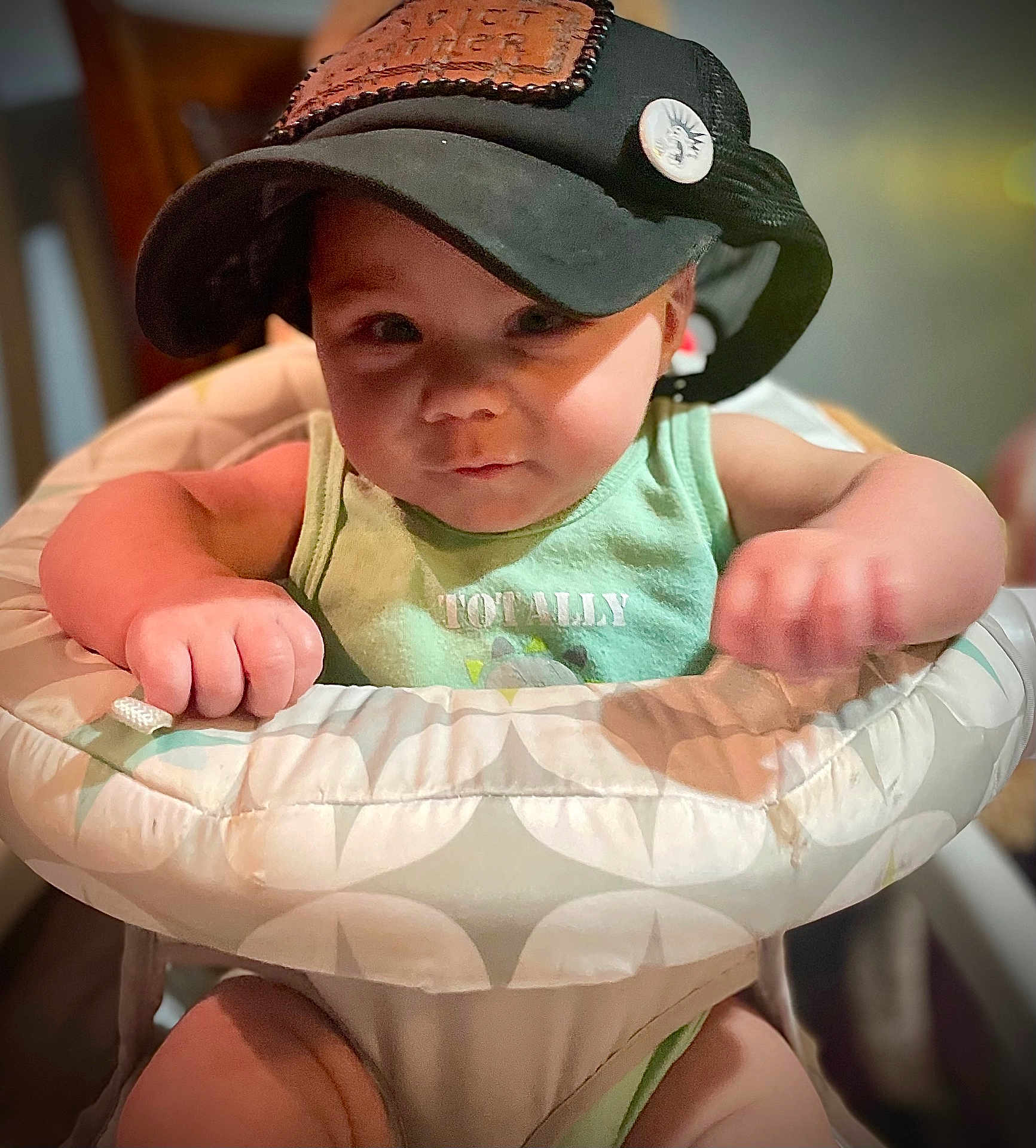 Victor joined the competition — help win amazing prizes! baby, infant, high_chair, cap, trucker_hat, green_tank_top, text_on_shirt, face, cheeks, hands, arms, tray, close_up, portrait, indoors, soft_lighting, button_pin, expression, background_blur, cute
