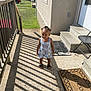 toddler, child, porch, sunlight, shadow, railing, steps, door, welcome_mat, outdoor, summer_clothing, sandals, house, concrete, grass, daylight, smiling, person, hair_buns, playful