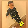 child, boy, portrait, standing, leaning, wall, indoor, black_outfit, yellow_shoes, necklace, fashion, hairstyle, face, person, carpet, baseboard, photograph, memory, date_text, text_overlay