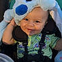 baby, child, smiling, hat, stuffed_animal, stroller, outdoor, sunlight, green_clothing, happy, blue, animal_costume, seatbelt, cute, infant, face, eyes, hand, playful, baby_toy