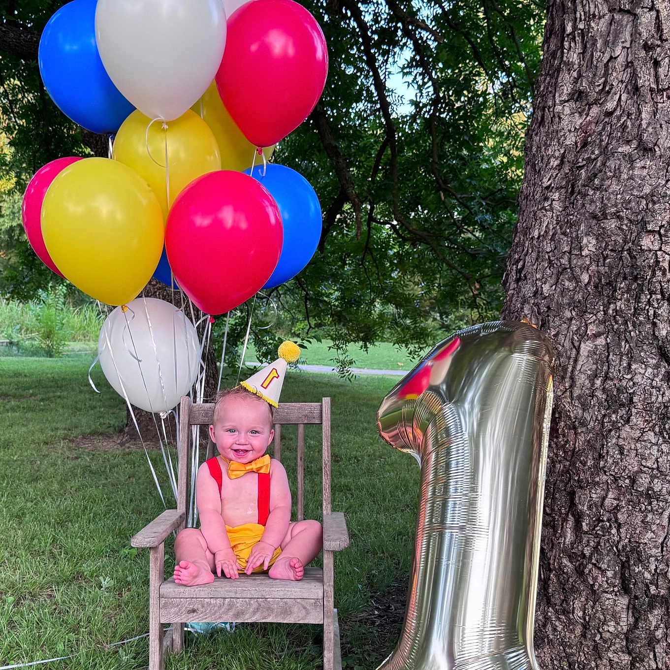Joseph is registered to the contest to win money with this photo: baby, birthday, balloons, outdoor, grass, tree, chair, party_hat, number_one, smiling, child, celebration, red, yellow, blue, green, wood, nature, fun, happy