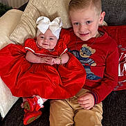 Santanna is registered to the contest to win money with this photo: baby, child, boy, couch, pillow, red_dress, white_bow, smile, face, sitting, indoor, happy, clothing, footwear, sibling, cute, holiday, portrait, home, furniture