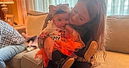 Kylie is registered to the contest to win money with this photo: baby, child, woman, couch, indoor, curtains, lamp, window, clothing, skirt, bow, holding, seated, person, home, portrait, warm_light, smile, family, cozy