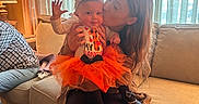 Kylie is registered to the contest to win money with this photo: affection, baby, bow, child, clothing, couch, cozy, curtains, dog_tail, furniture, home, indoor, kiss, lamp, orange_tutu, person, sitting, smiling, warm_lighting, woman