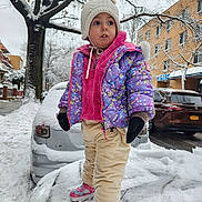 Norma is registered to the contest to win money with this photo: toddler, child, winter, snow, snowy_street, colorful_jacket, white_hat, mittens, snow_covered_car, residential_area, tree, sidewalk, curious_expression, outdoor, cold_weather, pants, sneakers, urban, daytime, person
