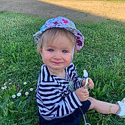 Eden a rejoint le concours — aidez-le/la à gagner de superbes lots ! toddler, child, grass, outdoor, hat, striped_shirt, blue_eyes, snack_pouch, drool, smile, sitting, sunlight, greenery, flowers, casual_clothing, playful, nature, cute, daylight, young_child