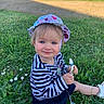 blue_eyes, casual_clothing, child, cute, daylight, drool, flowers, grass, greenery, hat, nature, outdoor, playful, sitting, smile, snack_pouch, striped_shirt, sunlight, toddler, young_child