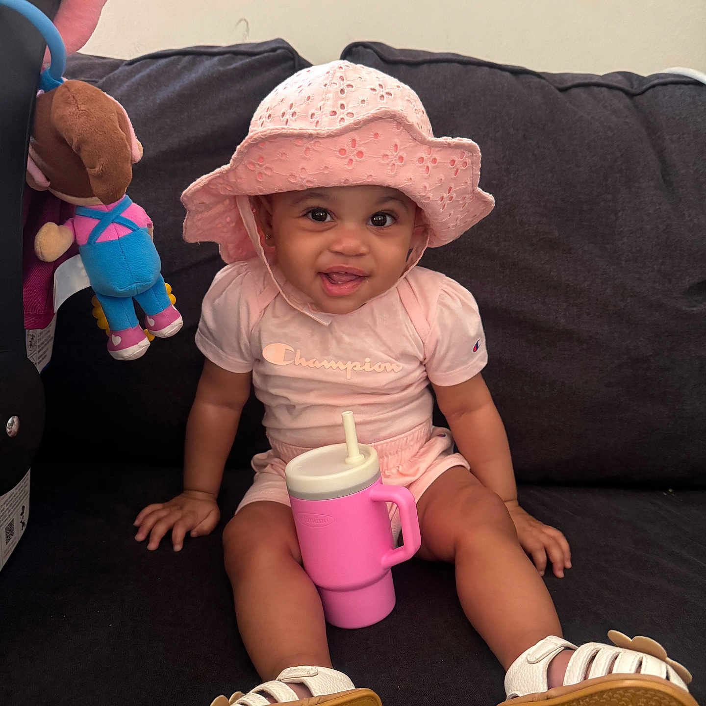 Paris is registered to the contest to win money with this photo: baby, child, pink_hat, pink_clothing, sippy_cup, smiling, sitting, couch, plush_toy, indoor, footwear, sandals, happy, cute, portrait, infant, toy, childhood, casual, home